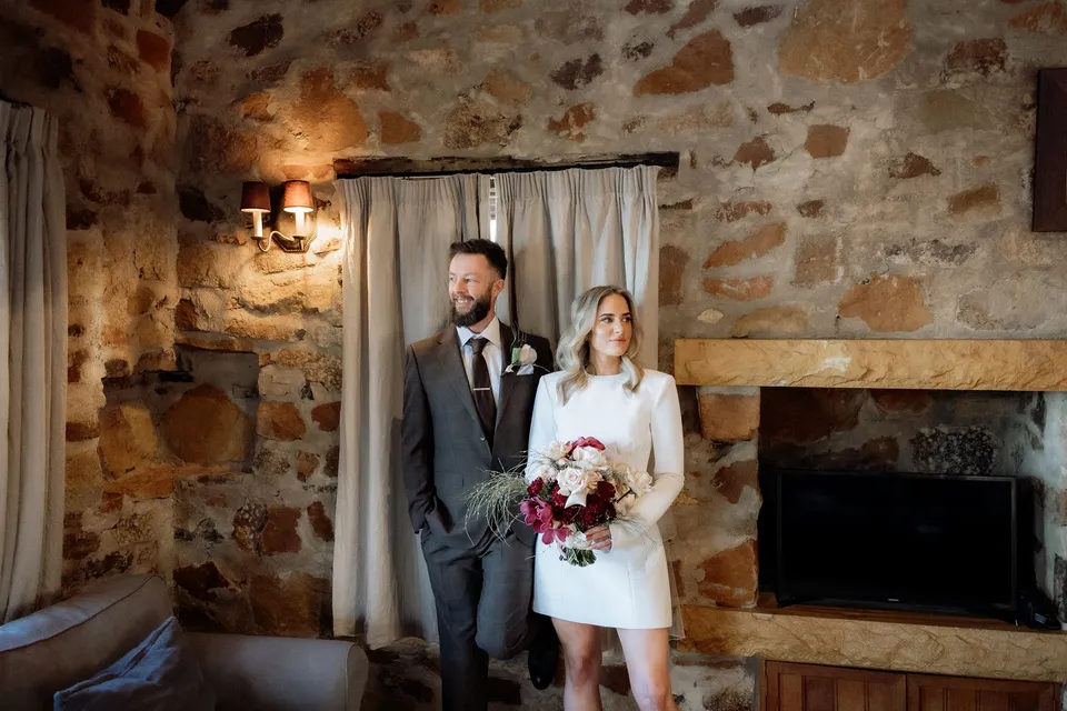 Kaitlyn and Russell standing beside the stone fireplace, with her bouquet by The Romantics and the timber ceiling above. At Piermont Retreat on Tasmania’s east coast, for an Elopement Collective elopement. Photograph by Omar of By The Wilde.