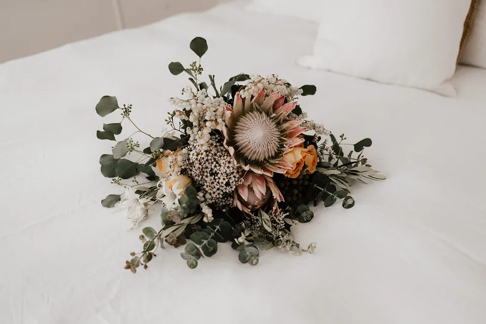 A bouquet by Fifth Avenue Florist with pink proteas, cream and apricot roses and eucalyptus resting on white bedding, at Grounds of Cabarita in Cabarita Beach, NSW, for an Elopement Collective elopement. Photo by Bulb Creative.