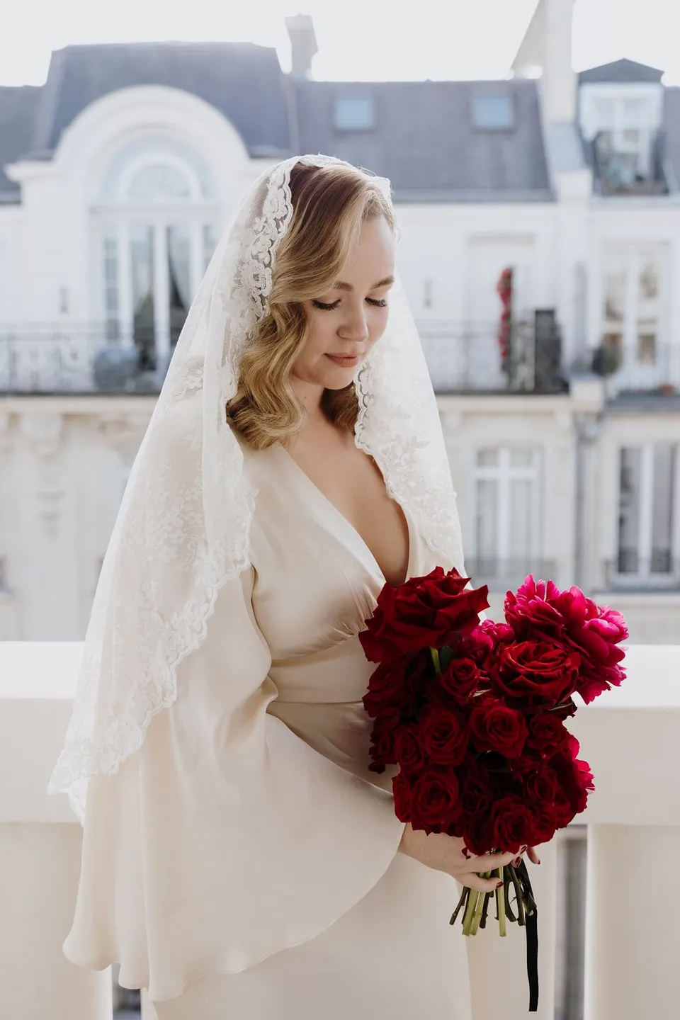 Kara in a white dress with long draped sleeves and a lace veil, holding her deep red bouquet by Ivy Bleu on the balcony, at Hotel Marignan Champs-Élysées in Paris, France, during an Elopement Collective elopement. Photograph by George Bowden.