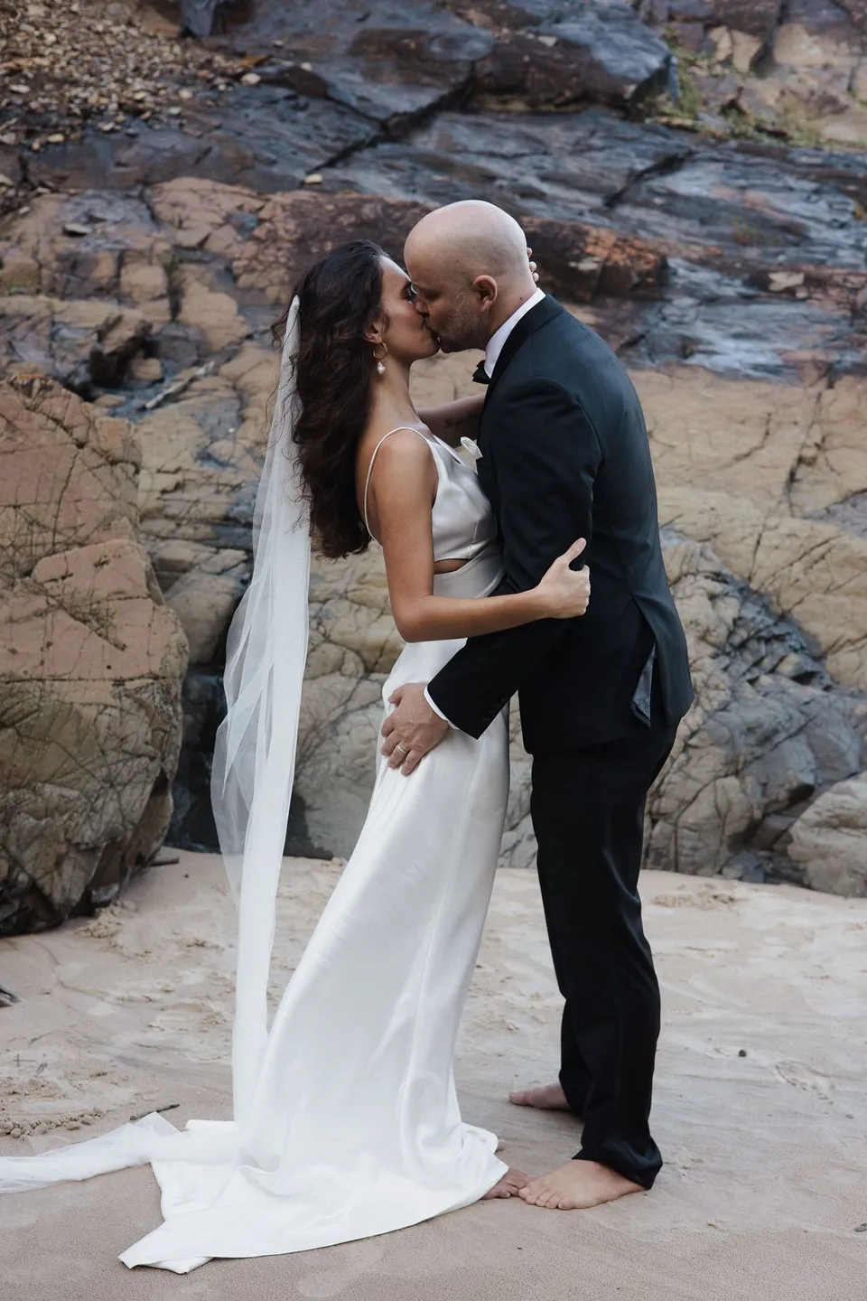 Rachel and Dane sharing a kiss on the sand beside the layered coastal rocks. At Whites Beach, Broken Head, near Byron Bay, for an Elopement Collective elopement. Photograph by Jason Lucas.