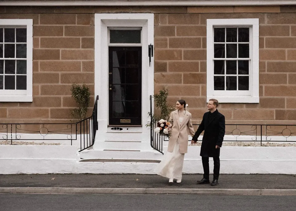 A couple hold hands outside a sandstone house with white steps and sash windows.