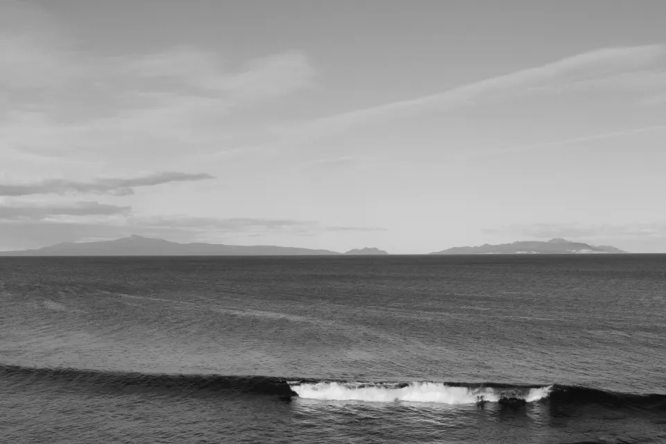 Black and white view of small waves breaking along the rocky shoreline, with distant hills across Great Oyster Bay, at Thalia Haven on Tasmania’s east coast, for an Elopement Collective elopement. Photograph by Bulb Creative.
