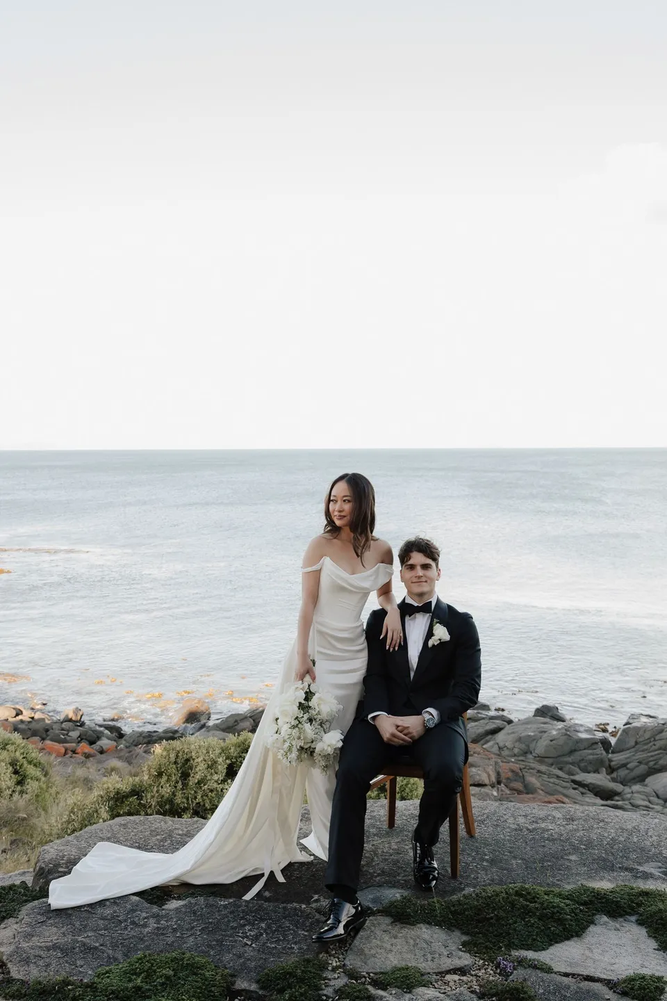 Clara standing beside Harry seated on a chair near the rocky shoreline, with her white bouquet by The Romantics and the sea behind them, at Thalia Haven on the East Coast of Tasmania, for an Elopement Collective elopement. Photograph by Bulb Creative.