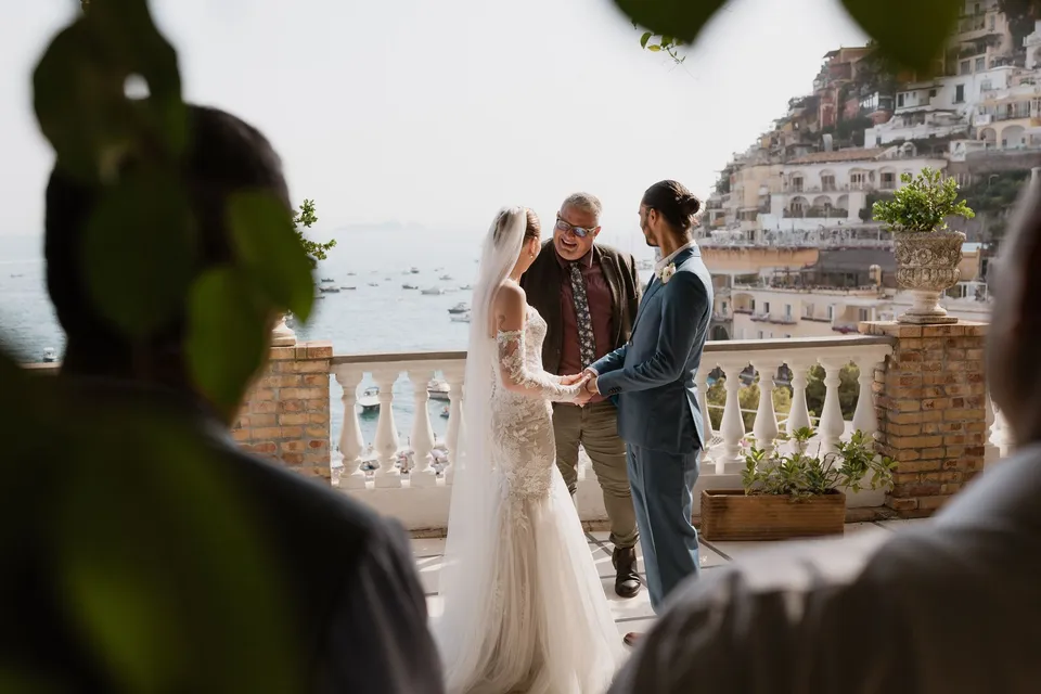 Skyler and Zach exchanging vows with celebrant Josh Withers on the terrace, seen past a seated guest, at Hotel Buca di Bacco in Positano, Amalfi Coast, Italy, for an Elopement Collective elopement. Photograph by Jason Corroto.