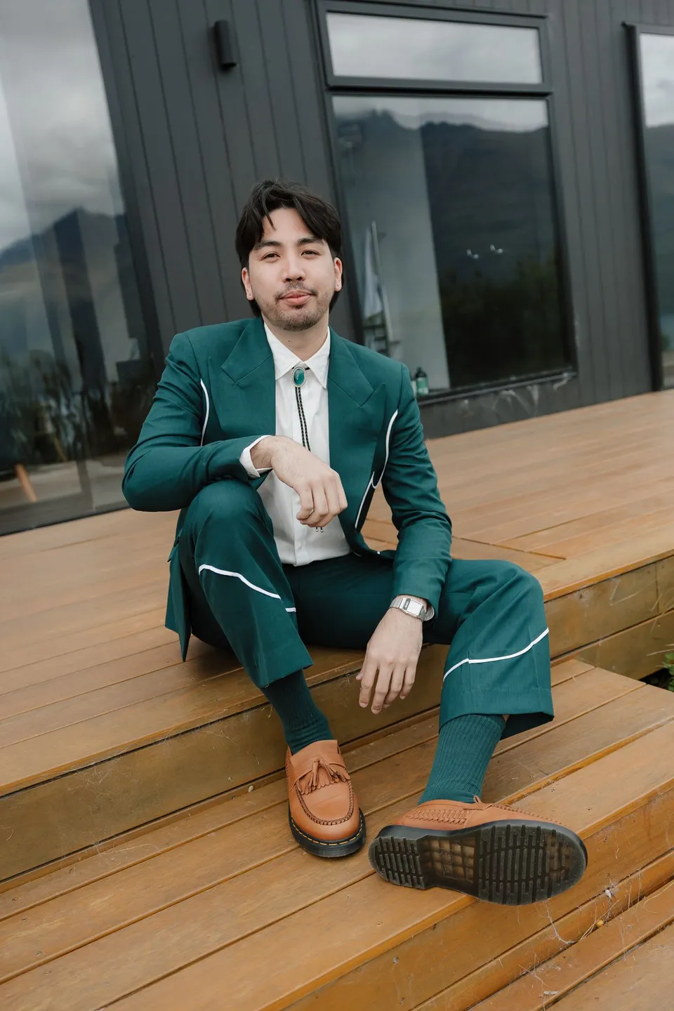 Adrian sitting on the timber deck in his green suit and tan loafers, at their alpine Airbnb in Ben Lomond, Queenstown, New Zealand, for an Elopement Collective Queenstown elopement. Photograph by Bulb Creative.