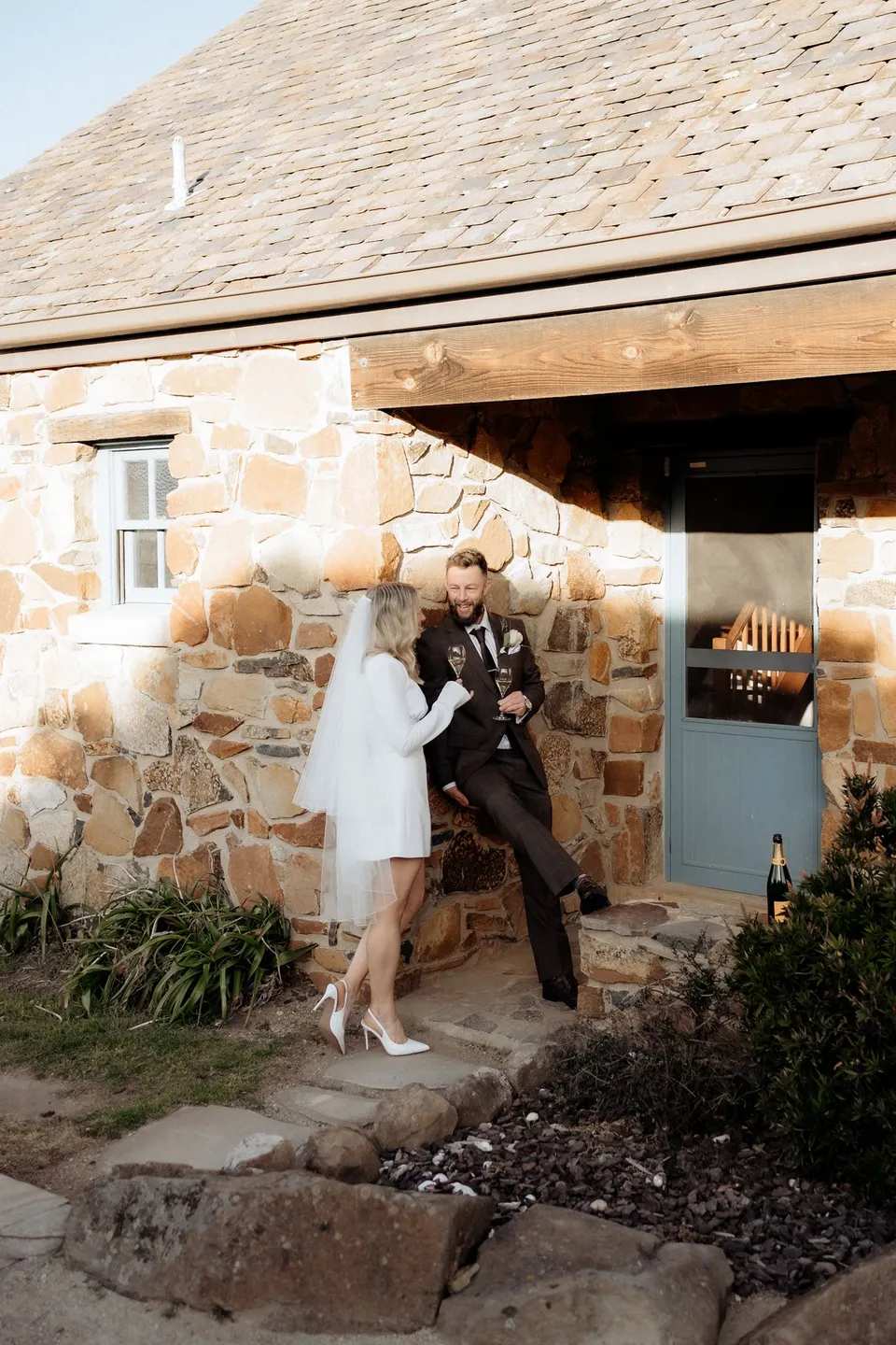 Kaitlyn and Russell stepping out of their stone cottage together, smiling in the sunshine. At Piermont Retreat on Tasmania’s east coast, for an Elopement Collective elopement. Photograph by Omar of By The Wilde.