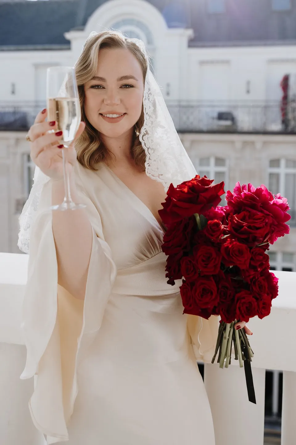 Kara smiling and raising a glass on the balcony, with her red bouquet by Ivy Bleu in her other hand, at Hotel Marignan Champs-Élysées in Paris, France, during an Elopement Collective elopement. Photograph by George Bowden.