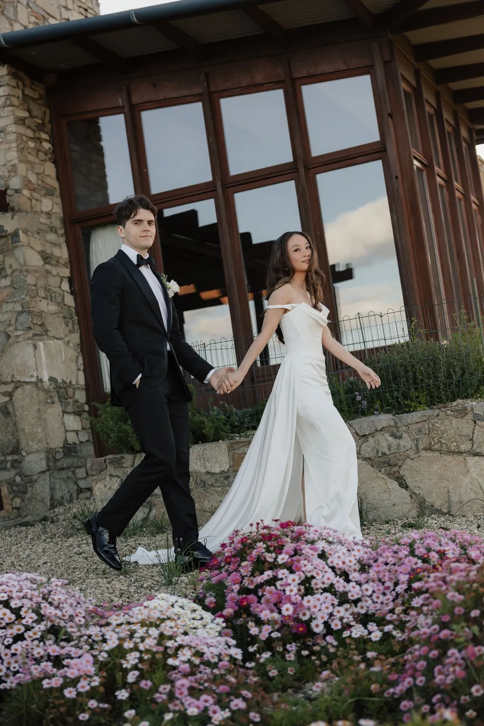 Clara and Harry walking hand in hand past the stone house and a bed of pink daisies, at Thalia Haven on the East Coast of Tasmania, for an Elopement Collective elopement. Photograph by Bulb Creative.