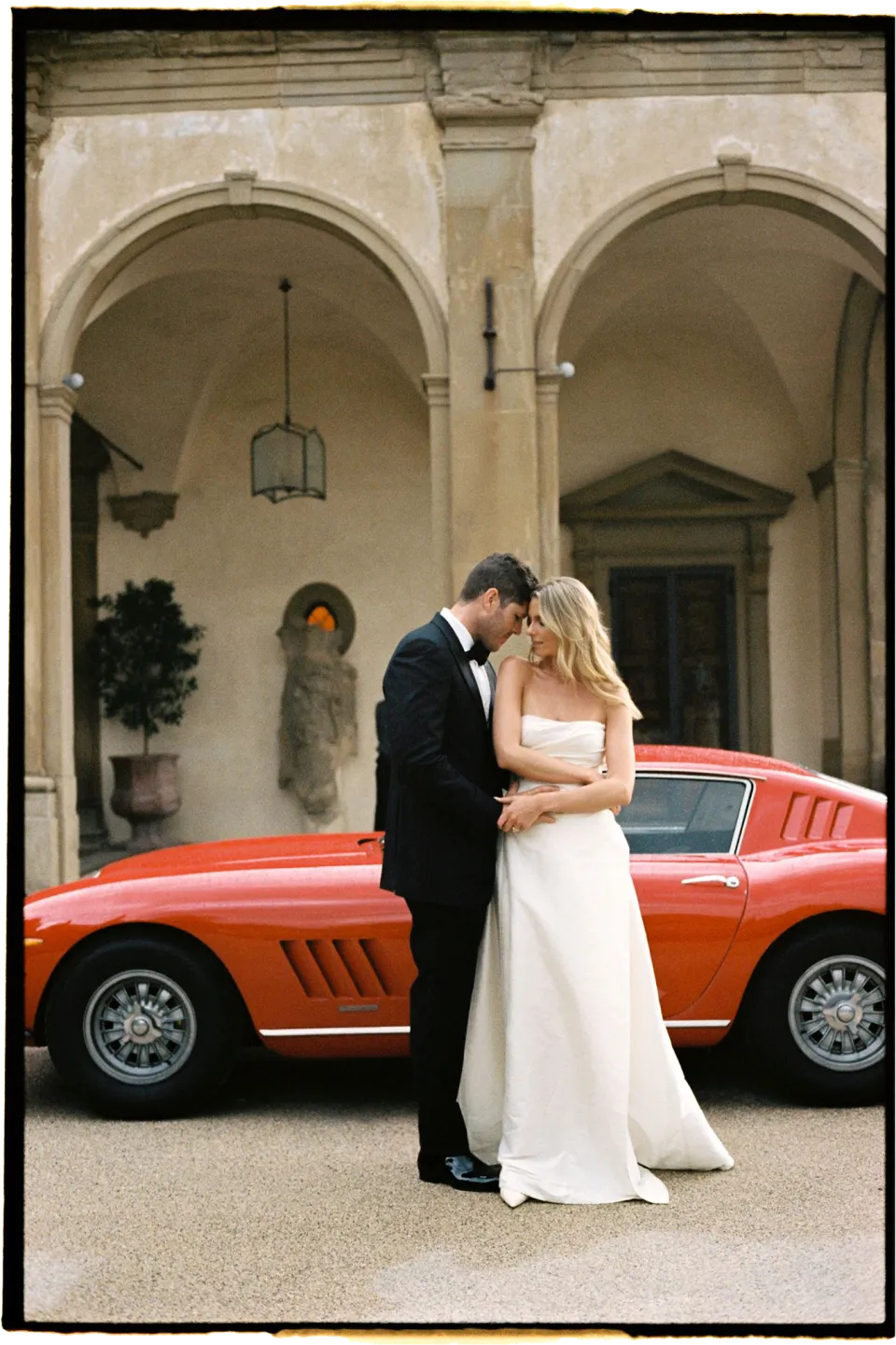 Matthew kissing Holly beside a red vintage sports car in the arcaded courtyard, at Villa San Michele in Fiesole above Florence, Tuscany, Italy, for an Elopement Collective elopement. Film photograph by Khara from House of Lucie.