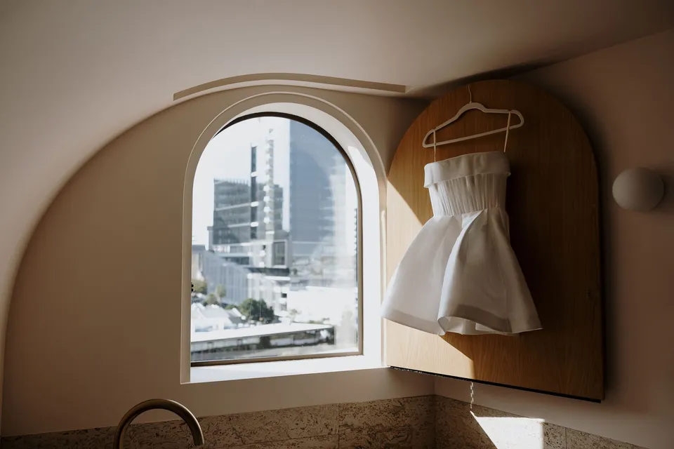 Danielle’s short white dress hanging beside an arched window overlooking Brisbane, during Danielle and Adam’s Elopement Collective elopement at The Calile Hotel in Fortitude Valley, Brisbane.
