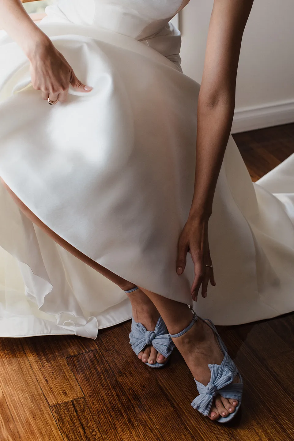 Meg lifting her wedding dress to fasten a pale blue bridal shoe on the timber floor. At The Keep on Tasmania’s east coast, for an Elopement Collective elopement. Photograph by Jenna Agius.