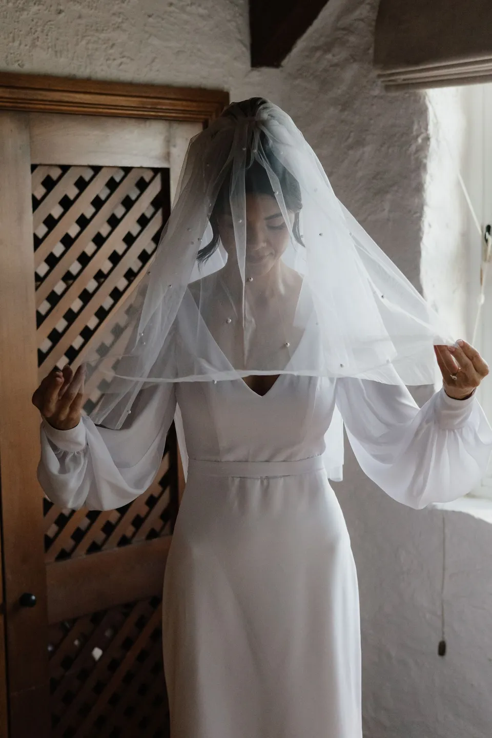 Renee lifting the edges of her veil in front of a timber fireplace surround inside the cottage. At Piermont Retreat on Tasmania’s east coast, for an Elopement Collective elopement. Photograph by Bulb Creative.