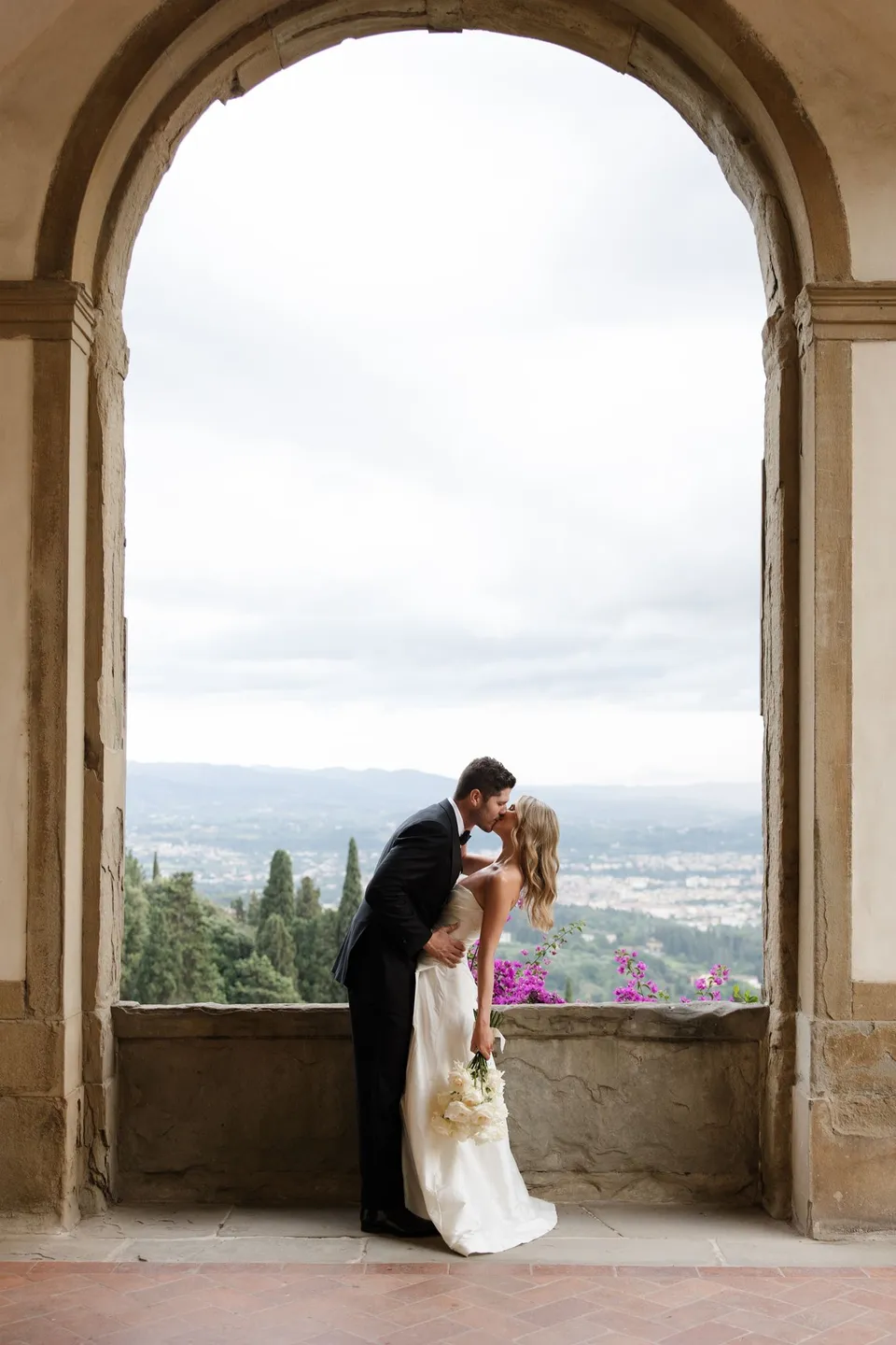 Holly and Matthew kissing beneath a stone arch, with her white bouquet by Le Francesche Flower Designers and Florence beyond, at Villa San Michele in Fiesole above Florence, Tuscany, Italy, for an Elopement Collective elopement. Digital photograph by Khara from House of Lucie.