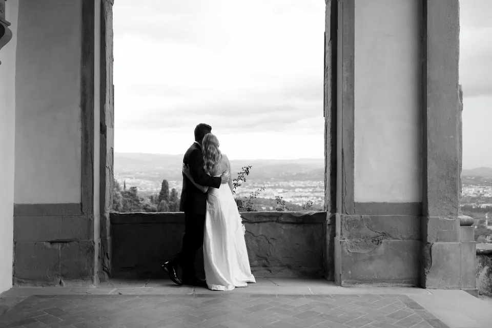 Black-and-white wide photograph of Holly and Matthew embracing beneath an arch in the villa loggia, at Villa San Michele in Fiesole above Florence, Tuscany, Italy, for an Elopement Collective elopement. Digital photograph by Khara from House of Lucie.