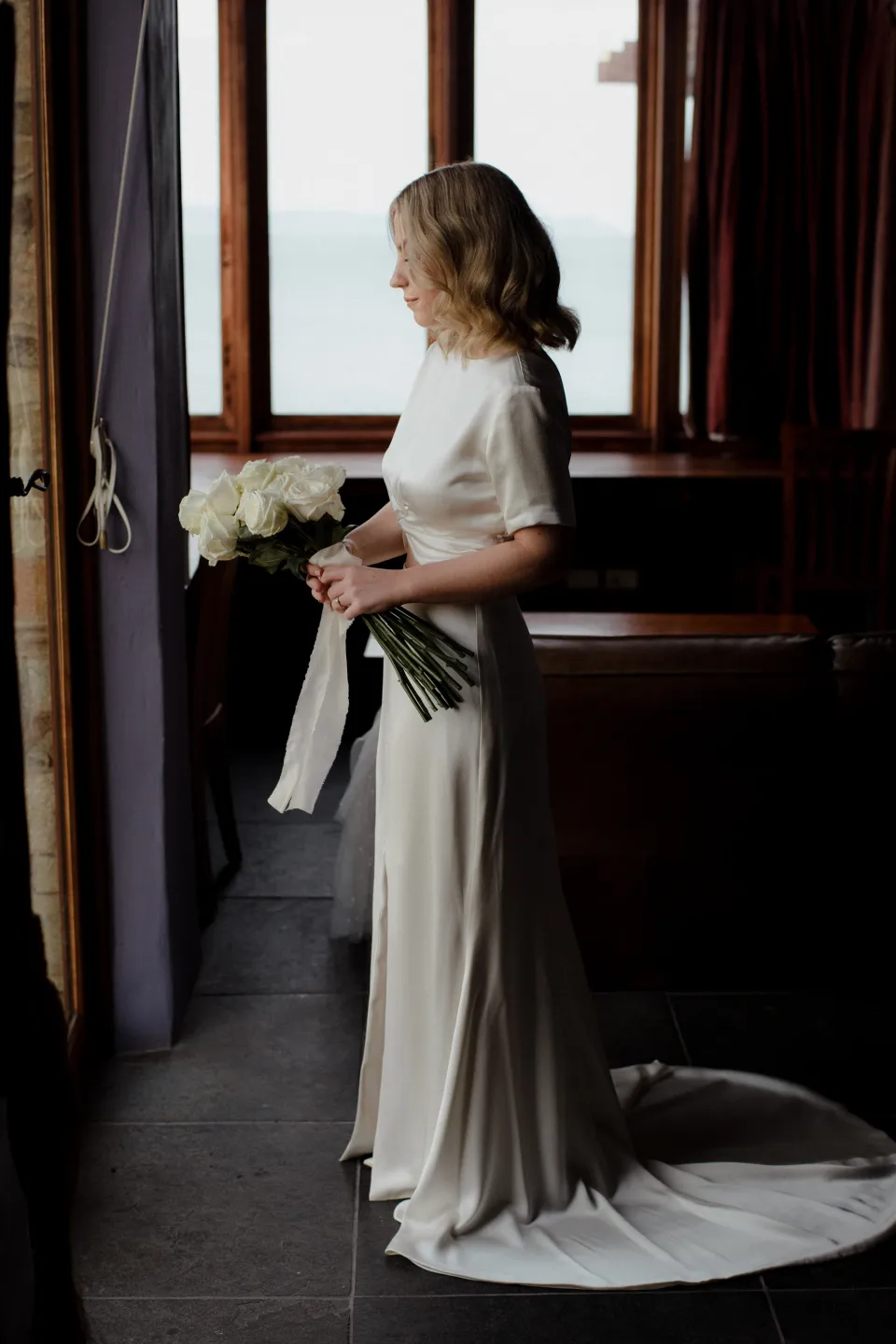 Jemmah looking at her bouquet by The Romantics beside the window, at Thalia Haven on the East Coast of Tasmania, Australia, for an Elopement Collective elopement. Photograph by Joey Willis.