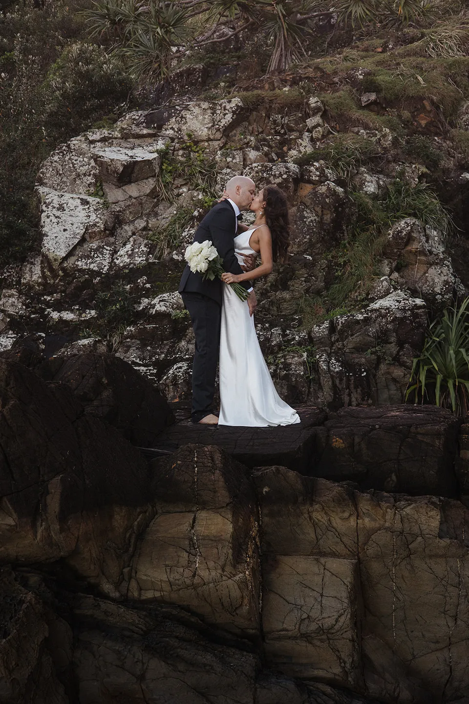 Rachel and Dane standing close together on a rocky ledge beside the beach. At Whites Beach, Broken Head, near Byron Bay, for an Elopement Collective elopement. Photograph by Jason Lucas.