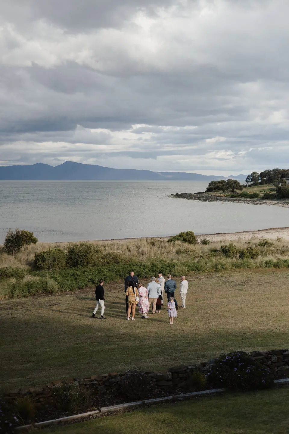 Guests walking across the lawn towards the ceremony, with the shoreline and cloud-covered hills beyond. At Piermont Retreat on Tasmania’s east coast, for an Elopement Collective elopement. Photograph by Bulb Creative.