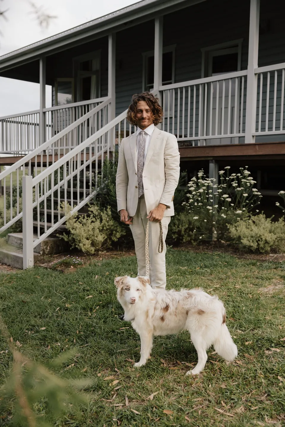 James in a cream suit standing beside a small white dog at the cottage steps. At Cudgera Creek in northern New South Wales, for an Elopement Collective elopement. Photograph by Bulb Creative.