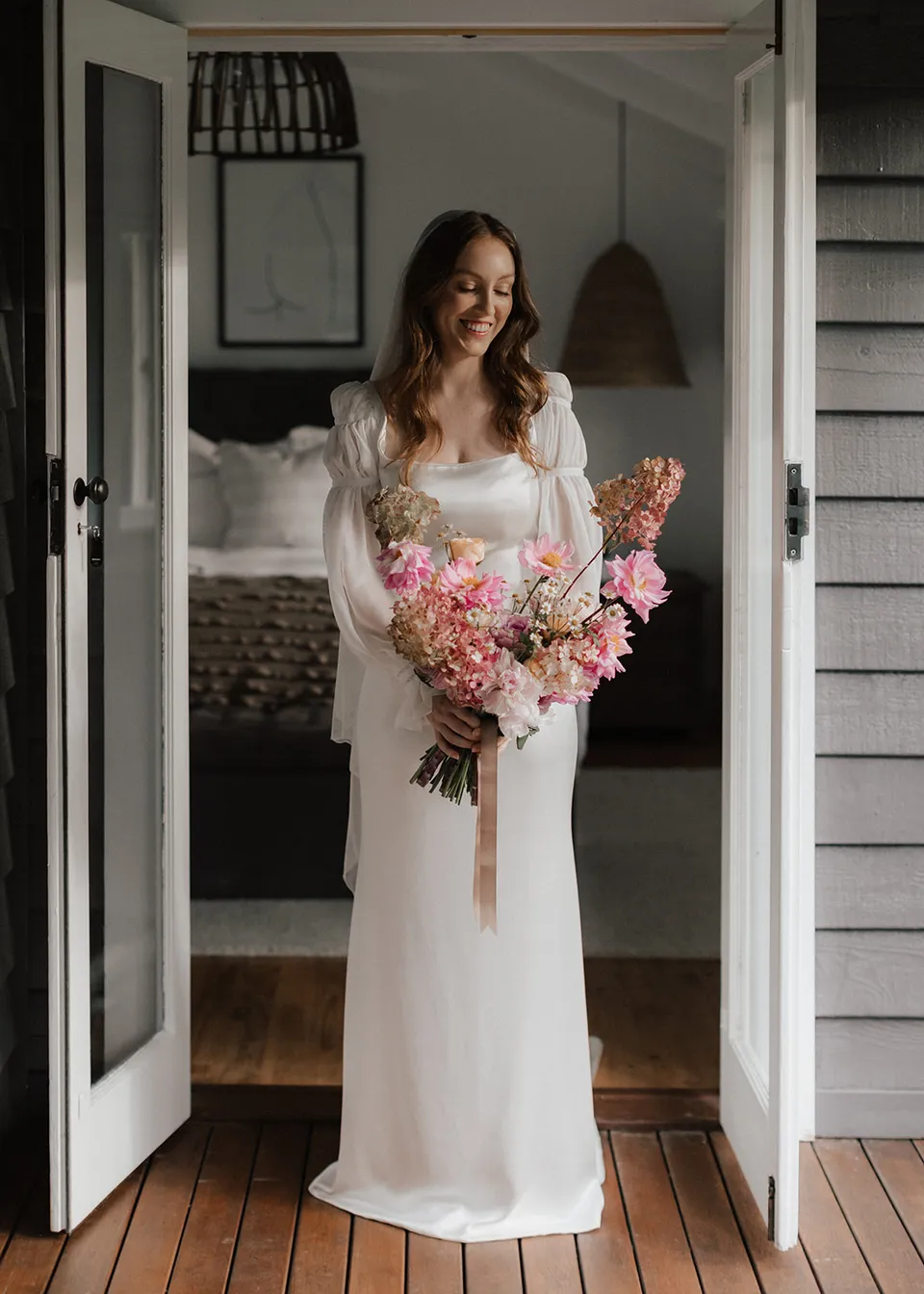Full-length portrait of Elyse holding her bouquet in the bedroom doorway, with flowers by The Perfect Party Co. At Fig Tree Villa in Ewingsdale, Byron Bay, for an Elopement Collective elopement. Photograph by Bulb Creative.