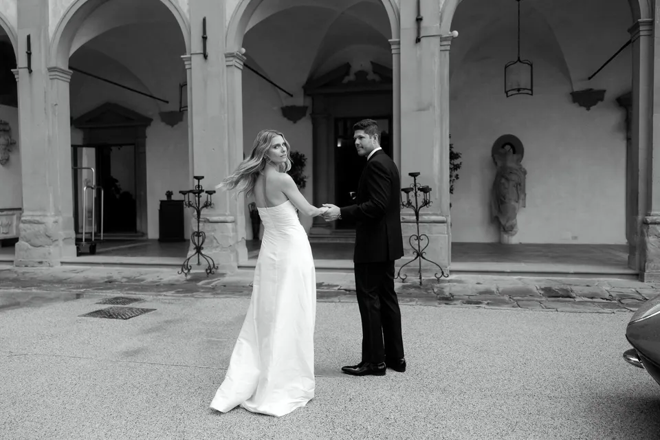 Black-and-white photograph of Holly and Matthew walking hand in hand across the arcaded courtyard, at Villa San Michele in Fiesole above Florence, Tuscany, Italy, for an Elopement Collective elopement. Digital photograph by Khara from House of Lucie.