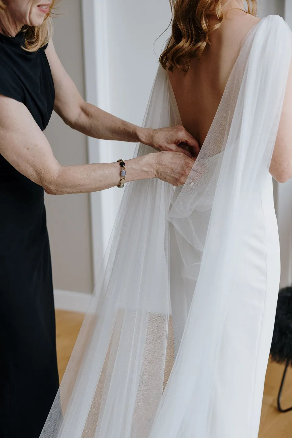 Neve having her white wedding dress fastened, with a sheer cape draped from her shoulders, at their Airbnb in Fernhill, Queenstown, New Zealand, for an Elopement Collective Queenstown elopement. Photo by George Bowden.