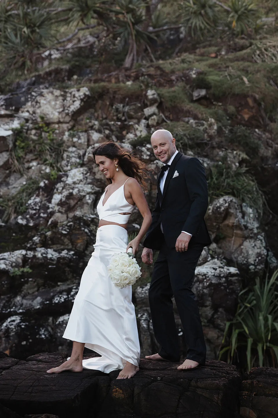 Rachel and Dane holding hands as they step carefully across the coastal rocks. At Whites Beach, Broken Head, near Byron Bay, for an Elopement Collective elopement. Photograph by Jason Lucas.