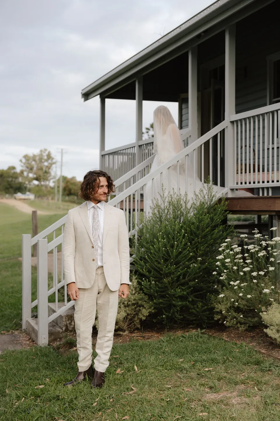 James waiting on the lawn as Bridget comes down the cottage stairs behind him. At Cudgera Creek in northern New South Wales, for an Elopement Collective elopement. Photograph by Bulb Creative.