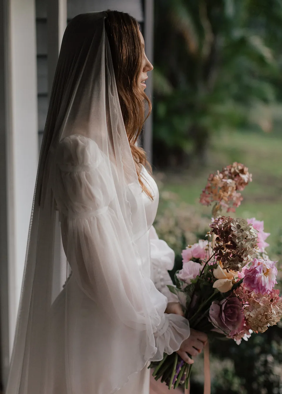 Side portrait of Elyse in her veil, holding flowers by the open door. At Fig Tree Villa in Ewingsdale, Byron Bay, for an Elopement Collective elopement. Photograph by Bulb Creative.