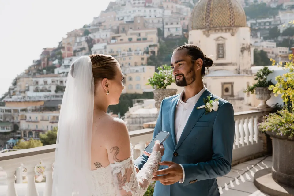Skyler reading her personal vows from her phone as Zach listens, with the tiled church dome and hillside houses behind them, at Hotel Buca di Bacco in Positano, Amalfi Coast, Italy, for an Elopement Collective elopement. Photograph by Jason Corroto.