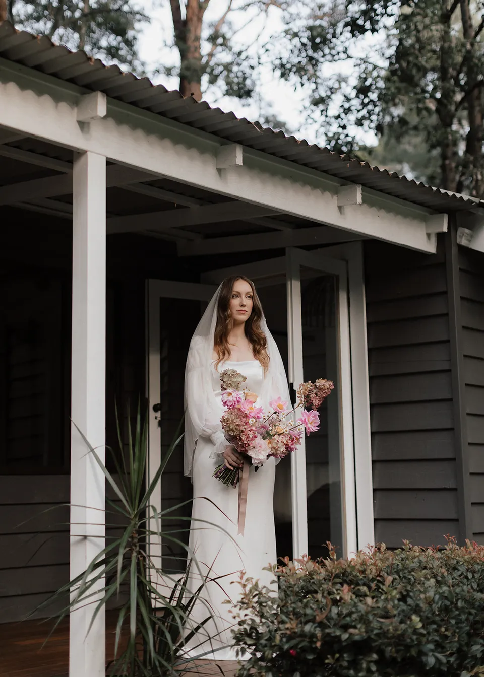 Elyse standing on the cottage verandah with her bouquet, with flowers by The Perfect Party Co. At Fig Tree Villa in Ewingsdale, Byron Bay, for an Elopement Collective elopement. Photograph by Bulb Creative.