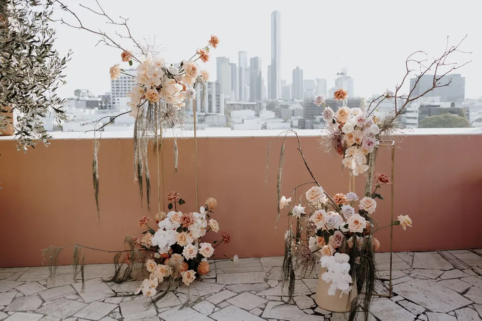 Peach and white ceremony flowers arranged among bare branches on a pink terrace overlooking Brisbane, during Danielle and Adam’s Elopement Collective elopement at The Calile Hotel in Fortitude Valley, Brisbane.