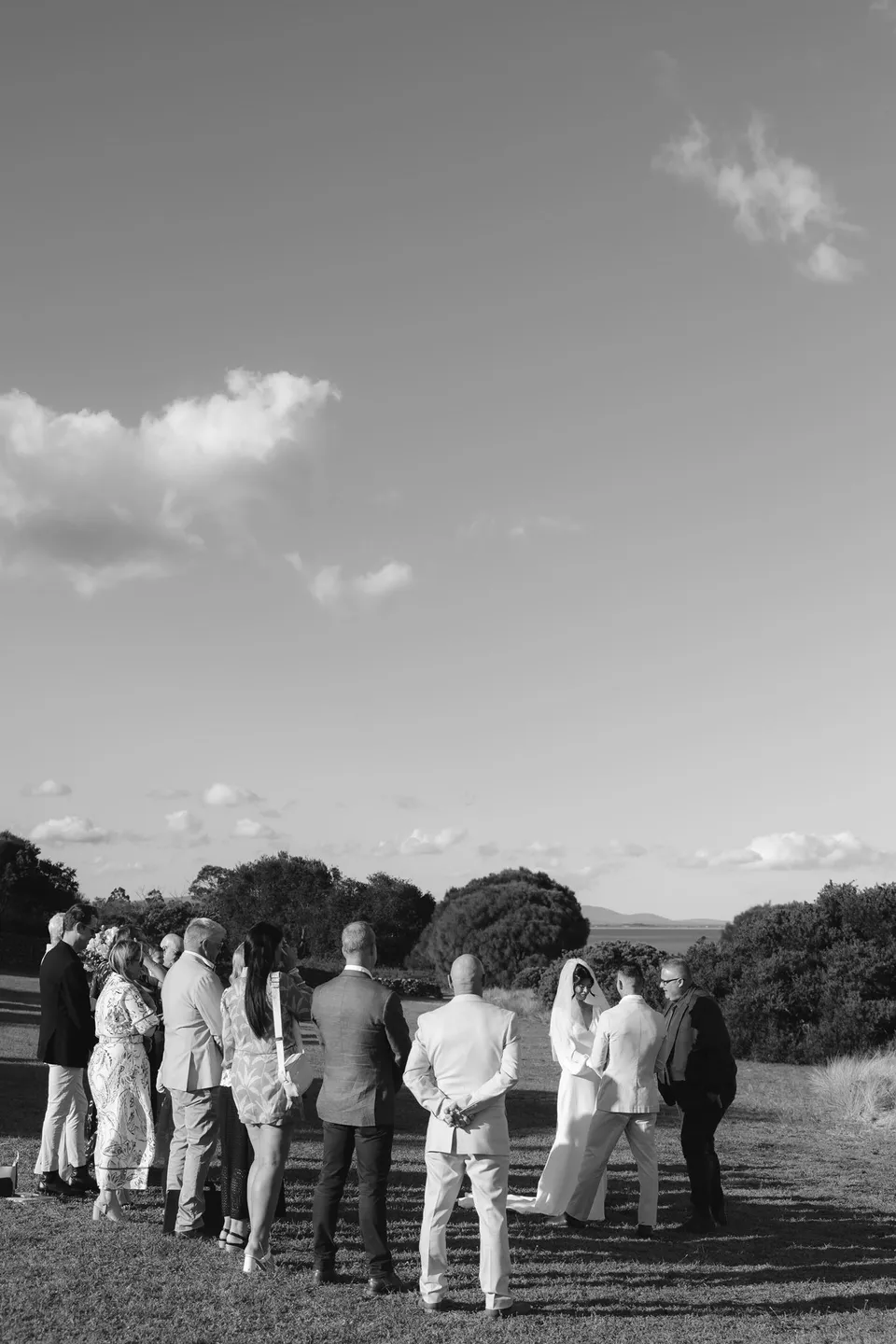 Black and white photograph of the small wedding gathering standing together on the coastal lawn. At Piermont Retreat on Tasmania’s east coast, for an Elopement Collective elopement. Photograph by Bulb Creative.