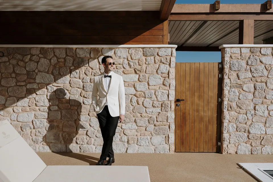 Brad wearing a white jacket, black bow tie and sunglasses beside a stone wall beneath the villa’s timber pergola, at Adriana Luxury Villas on Milos, Greece, during an Elopement Collective elopement. Photograph by George Bowden.