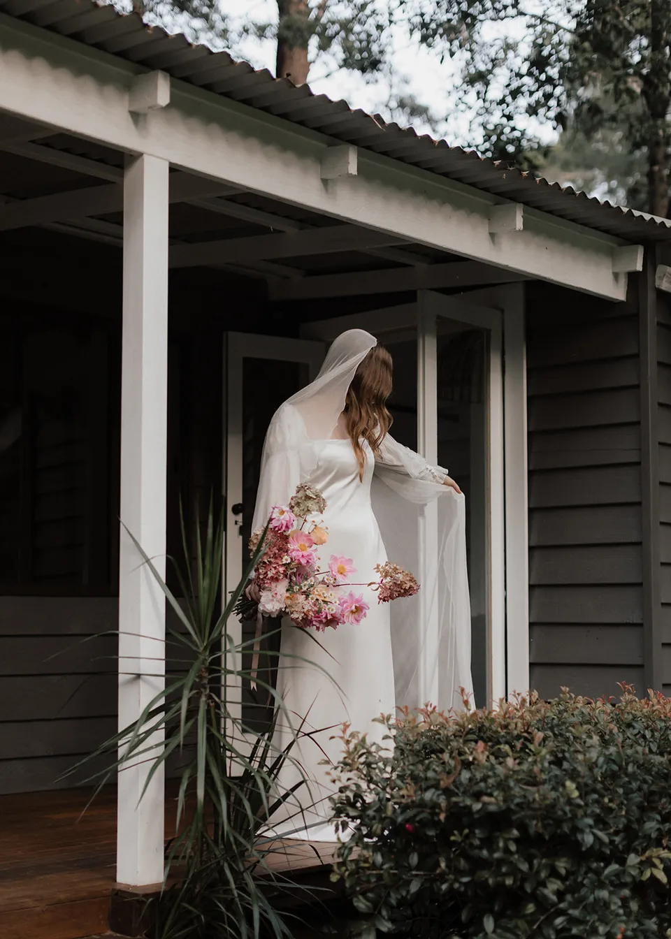 Elyse looking out from the verandah with flowers held at her side. At Fig Tree Villa in Ewingsdale, Byron Bay, for an Elopement Collective elopement. Photograph by Bulb Creative.
