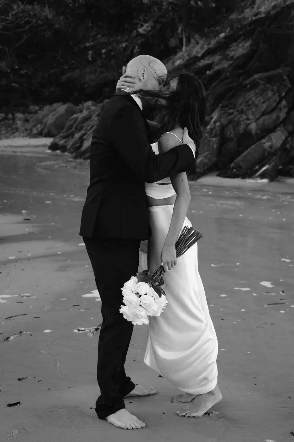 Black-and-white photograph of Rachel and Dane embracing on the sand. At Whites Beach, Broken Head, near Byron Bay, for an Elopement Collective elopement. Photograph by Jason Lucas.