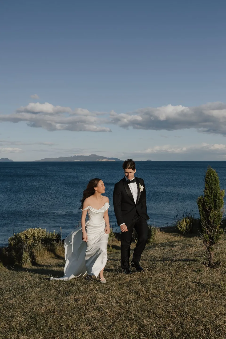 Clara and Harry walking together through golden grassland above the blue bay, at Thalia Haven on the East Coast of Tasmania, for an Elopement Collective elopement. Photograph by Bulb Creative.