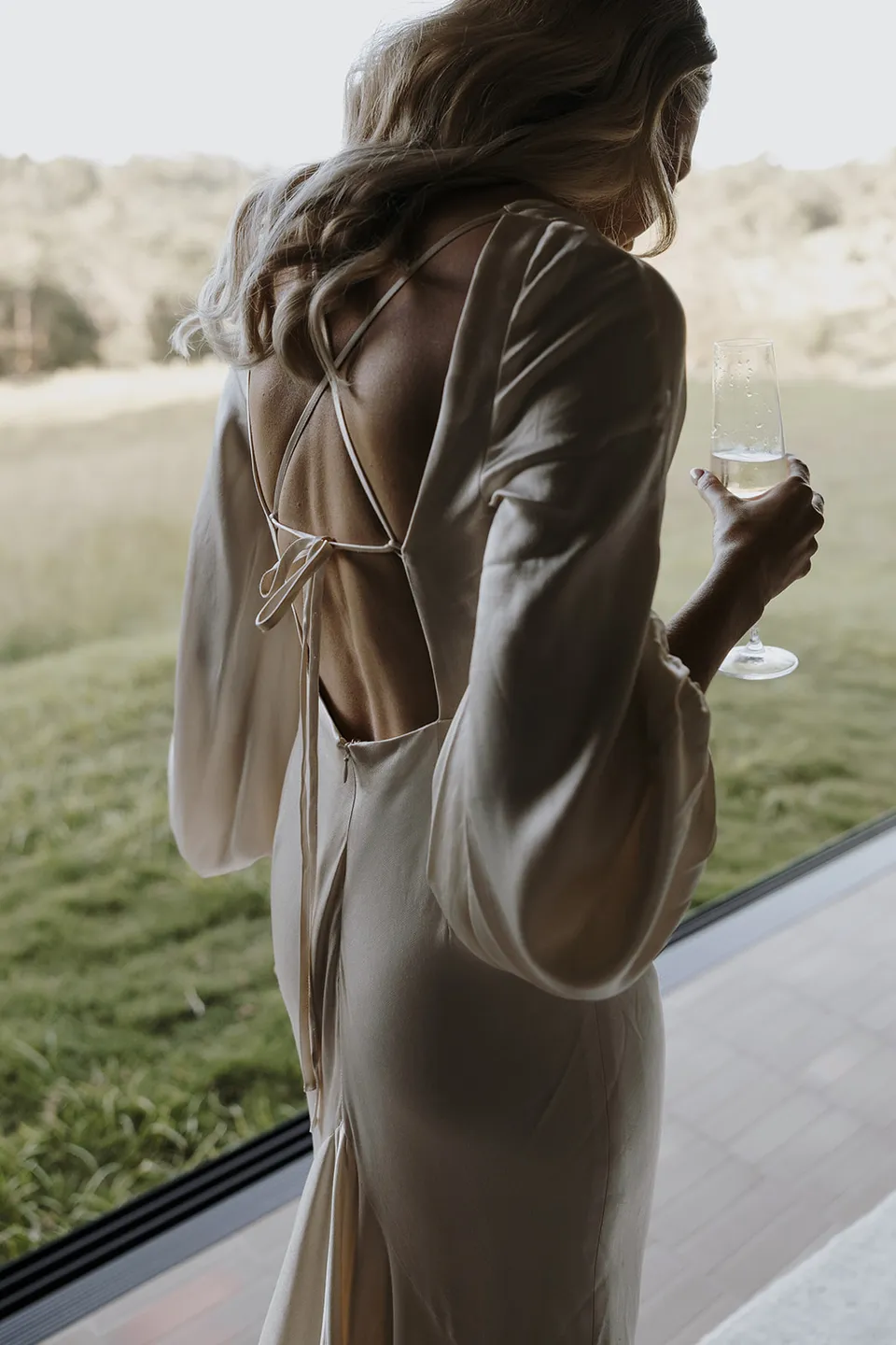 Gabrielle holding a glass of champagne beside the cabin window, showing her dress’s open back and tie detail at Roundhill Retreat in Obi Obi, Sunshine Coast Hinterland, for their Elopement Collective elopement.