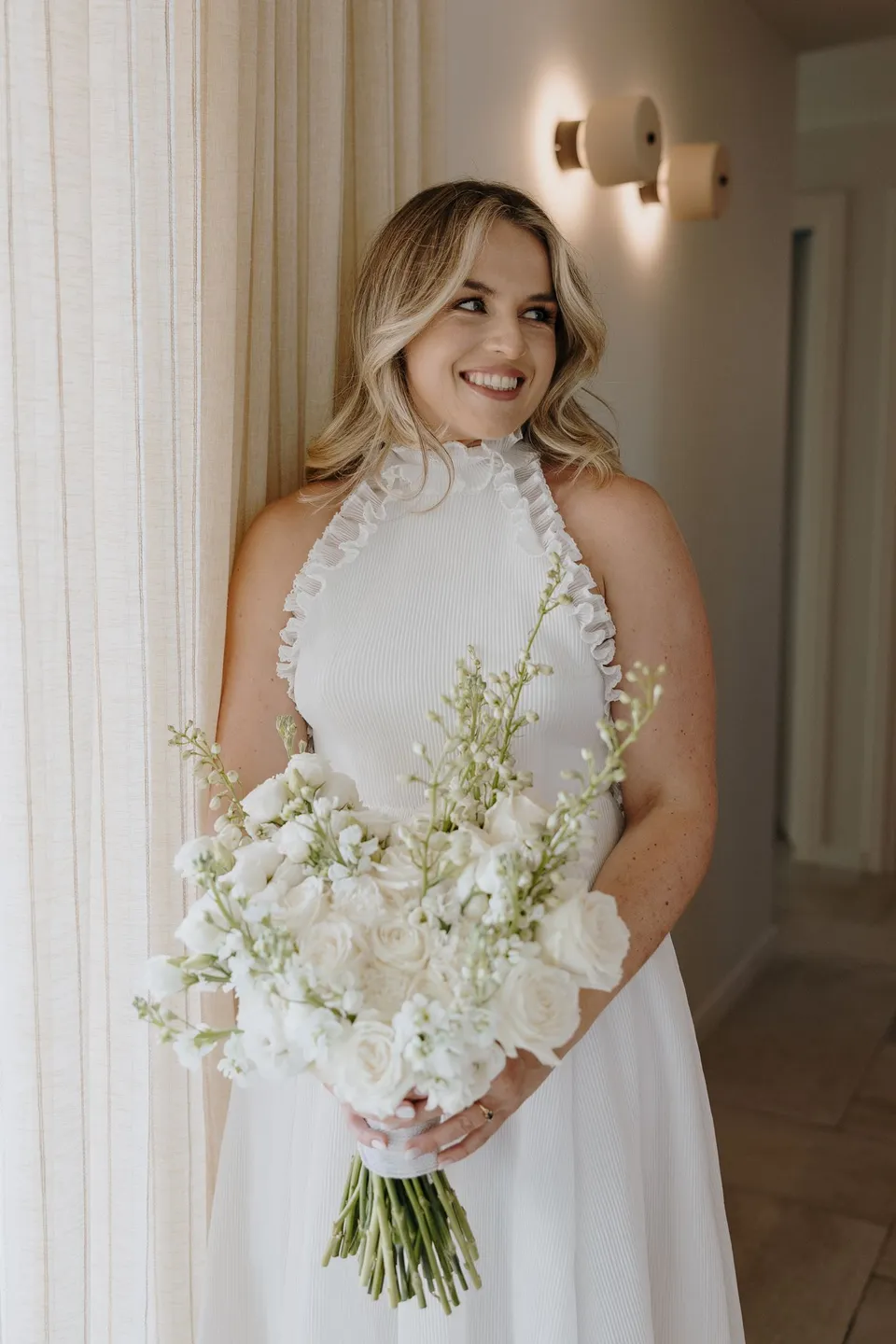 Georgina smiling in a villa doorway in her white halterneck dress, holding a white bouquet by Giasemi, at Adriana Luxury Villas on Milos, Greece, during an Elopement Collective elopement. Photograph by George Bowden.
