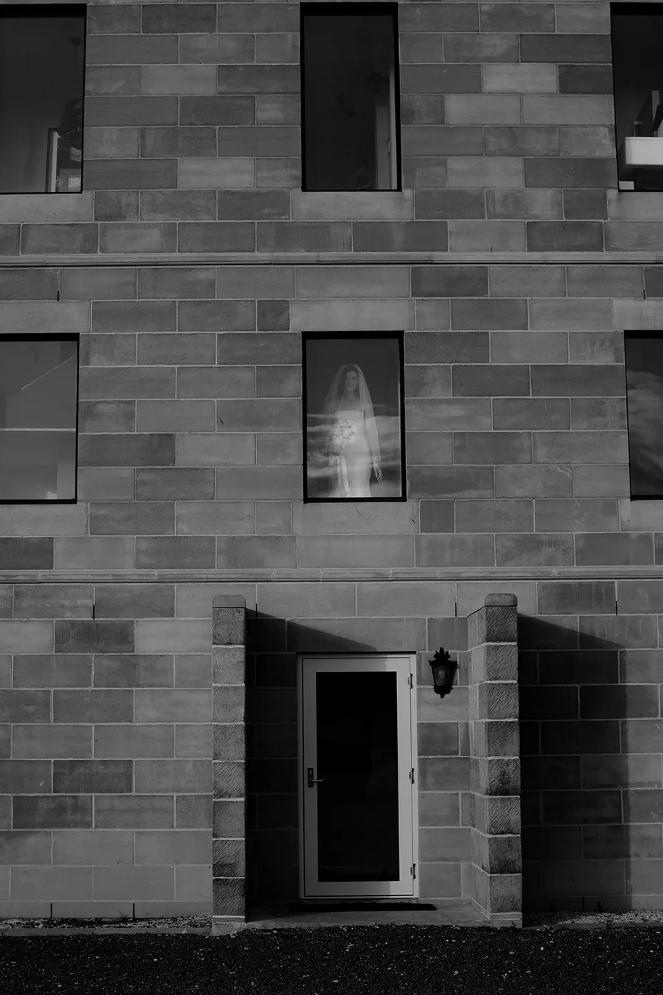 A bride visible through an upstairs window above The Keep’s open doorway, framed by the sandstone facade in black and white. At The Keep on Tasmania’s east coast, for an Elopement Collective elopement. Photograph by Jenna Agius.
