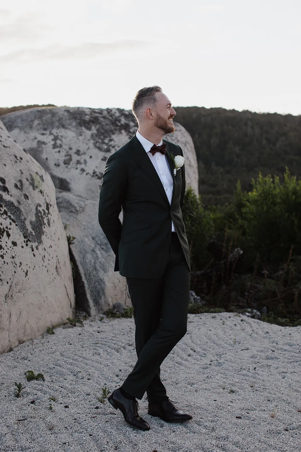 Ryan in his dark suit and bow tie, looking towards the hills beside a granite boulder. At The Keep on Tasmania’s east coast, for an Elopement Collective elopement. Photograph by Jenna Agius.
