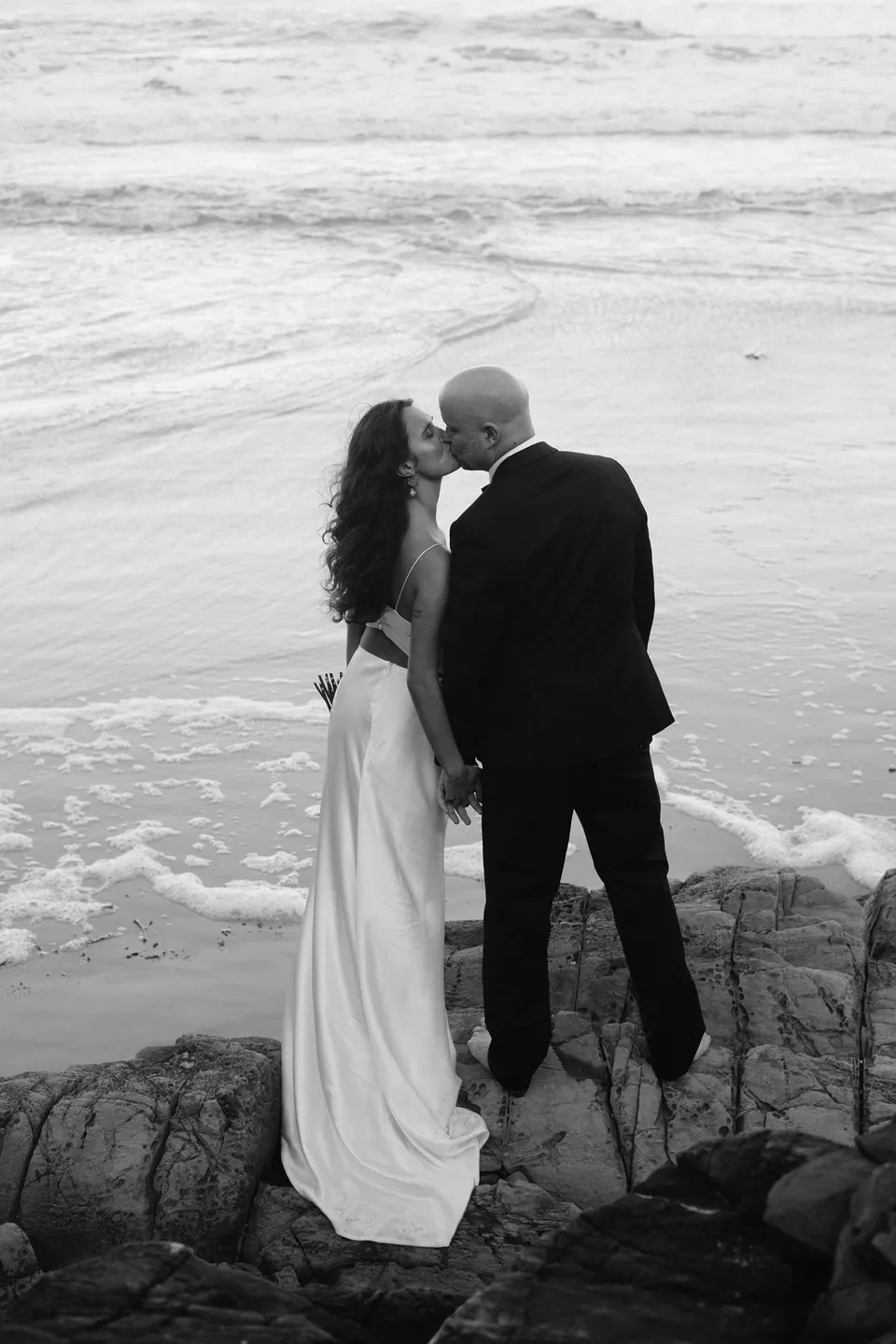 Black-and-white rear view of Rachel and Dane standing close on the rocks above the surf. At Whites Beach, Broken Head, near Byron Bay, for an Elopement Collective elopement. Photograph by Jason Lucas.