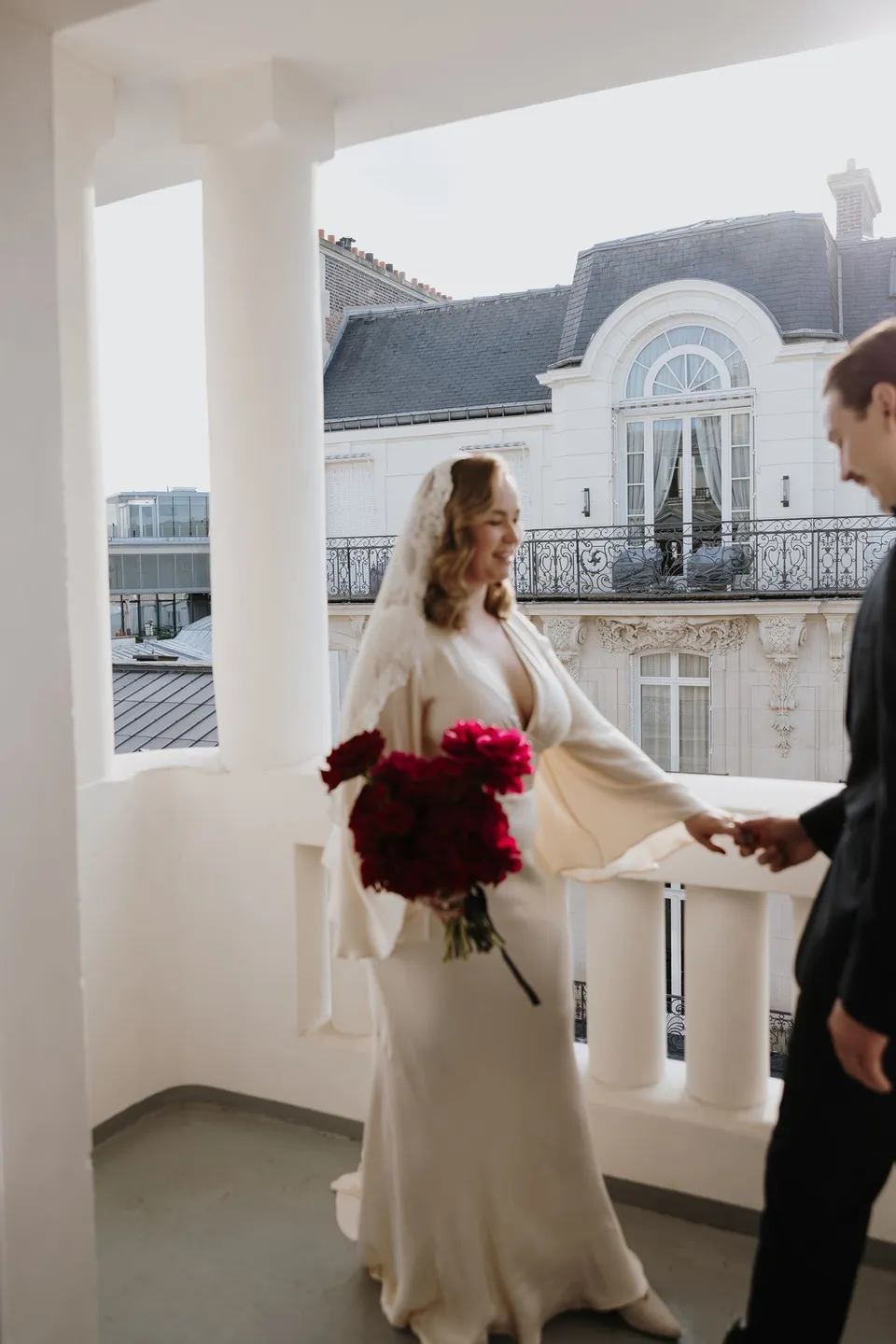 Kara and Samuel holding hands on the hotel balcony, with her red Ivy Bleu bouquet and his black tuxedo, at Hotel Marignan Champs-Élysées in Paris, France, during an Elopement Collective elopement. Photograph by George Bowden.