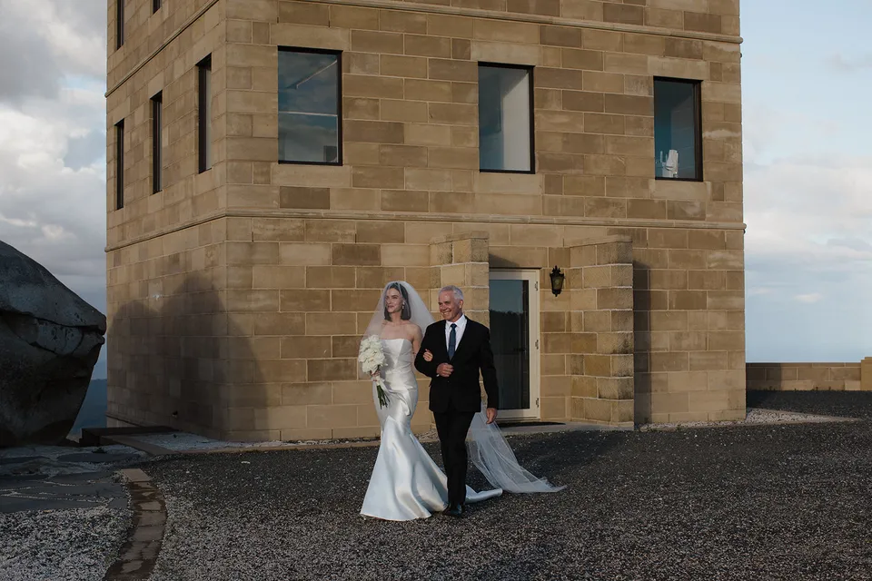 Meg walking arm in arm with a companion in front of The Keep’s sandstone tower. At The Keep on Tasmania’s east coast, for an Elopement Collective elopement. Photograph by Jenna Agius.