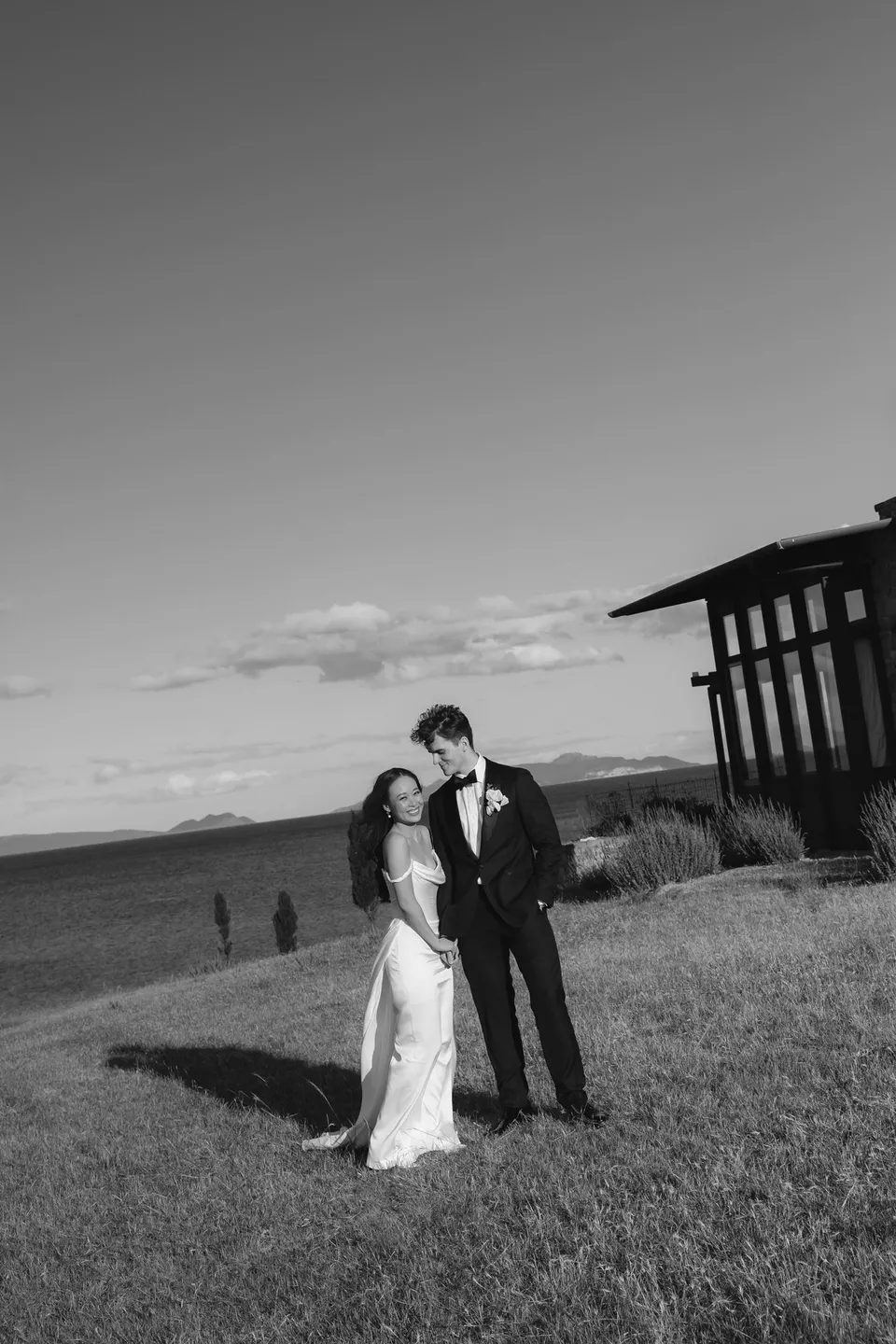 Black and white photograph of Clara and Harry standing close in coastal grassland beside the house, at Thalia Haven on the East Coast of Tasmania, for an Elopement Collective elopement. Photograph by Bulb Creative.