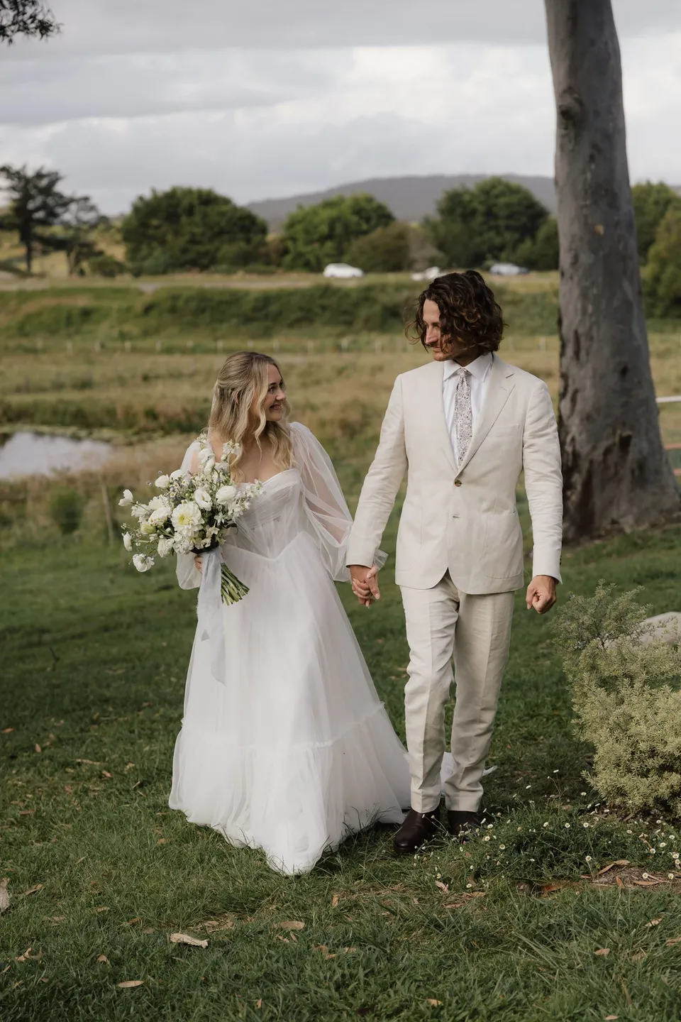 Bridget and James walking hand in hand across the lawn with rolling hills behind them. At Cudgera Creek in northern New South Wales, for an Elopement Collective elopement. Photograph by Bulb Creative.