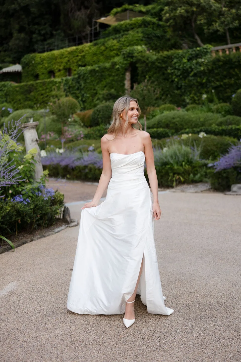 Holly holding the skirt of her white dress on a gravel path in the terraced garden, with bridal makeup by Shannon Hope, at Villa San Michele in Fiesole above Florence, Tuscany, Italy, for an Elopement Collective elopement. Digital photograph by Khara from House of Lucie.