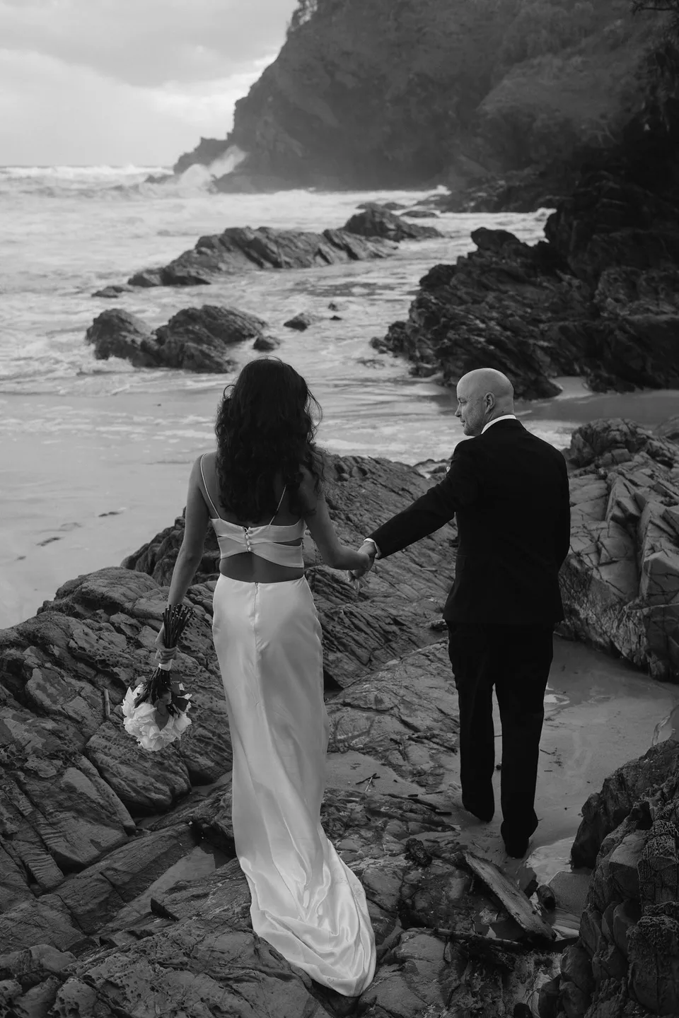 Black-and-white photograph of Rachel and Dane holding hands on the rocky shoreline. At Whites Beach, Broken Head, near Byron Bay, for an Elopement Collective elopement. Photograph by Jason Lucas.
