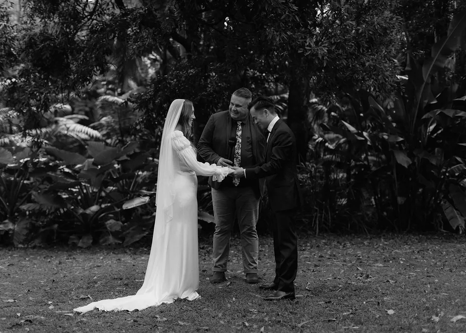 Black-and-white photograph of Elyse and Jonathan holding hands with celebrant Josh Withers beneath the trees. At Fig Tree Villa in Ewingsdale, Byron Bay, for an Elopement Collective elopement. Photograph by Bulb Creative.