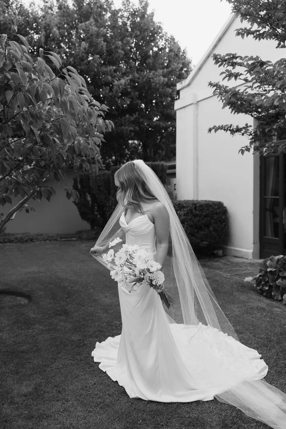 Black and white portrait of Tanesha holding her bouquet by The Romantics, with her veil and dress trailing across the garden lawn, at Islington Hotel in South Hobart, Tasmania, Australia, for an Elopement Collective summer elopement. Photograph by George Bowden.