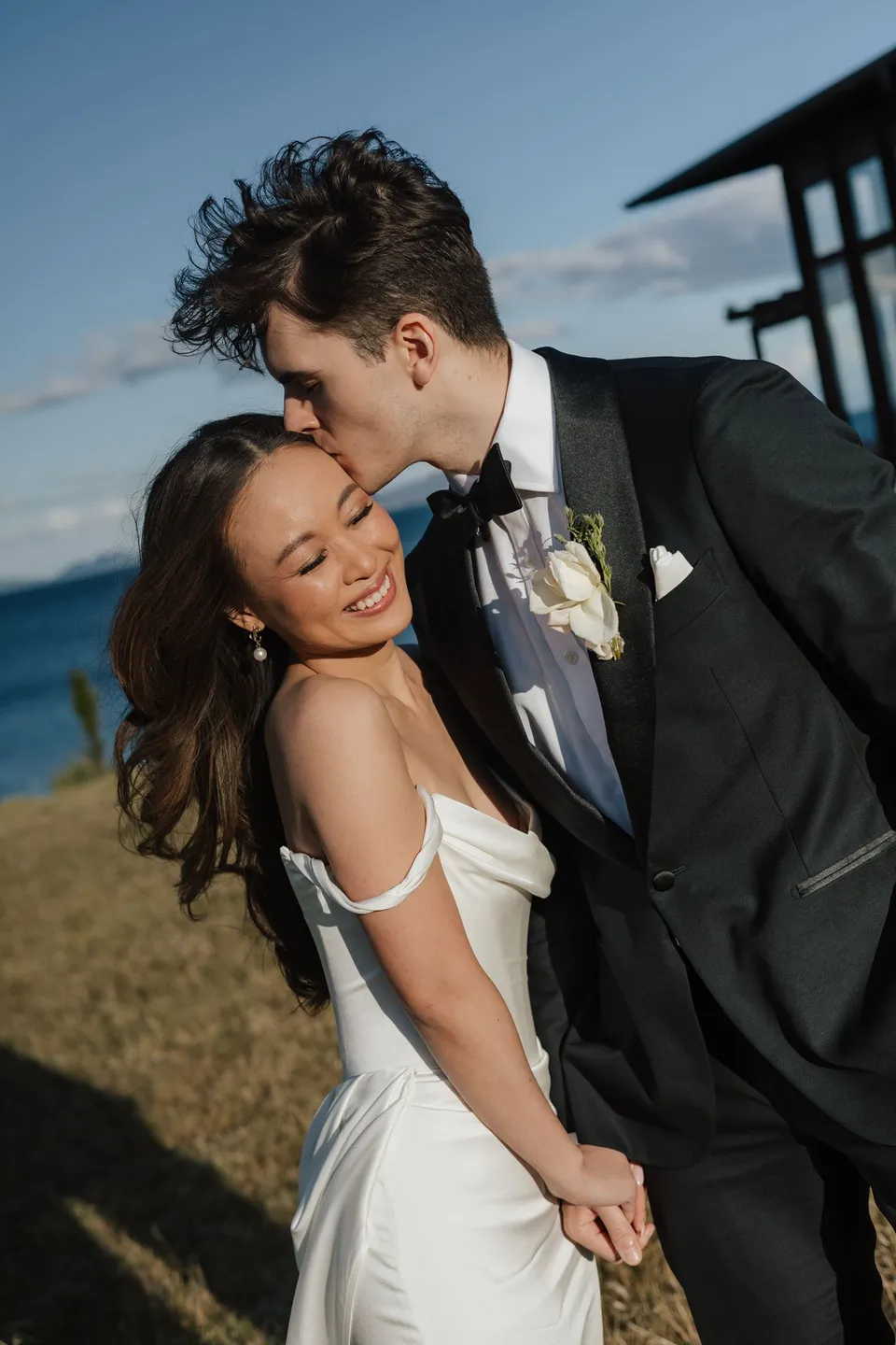 Harry kissing Clara’s forehead as she smiles and holds his hand, with her hair moving in the breeze, at Thalia Haven on the East Coast of Tasmania, for an Elopement Collective elopement. Photograph by Bulb Creative.